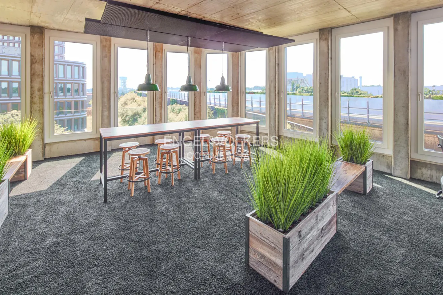 A modern office space with a long wooden table, stools, and large windows. Green plants in wooden planters add a touch of nature.