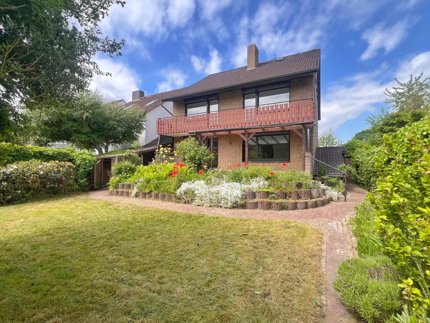 Two-story brick house with a brown balcony, surrounded by a green lawn and flower garden under a blue sky.