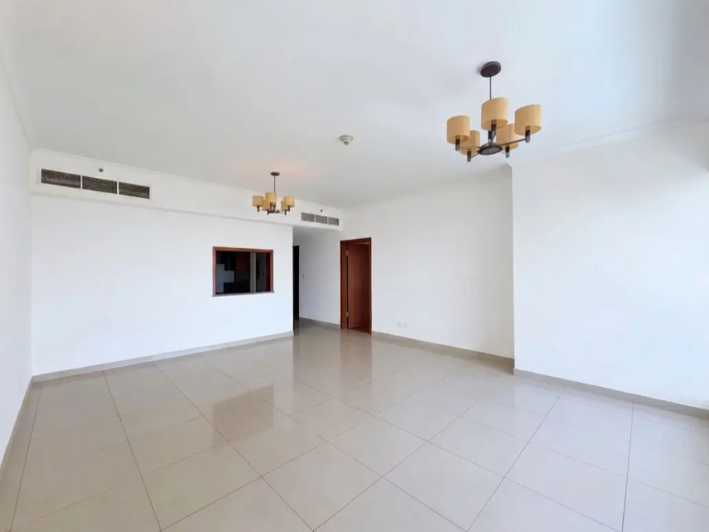 Bright, empty room with white walls, beige tile floor, and two chandeliers. A small window and a doorway are visible.