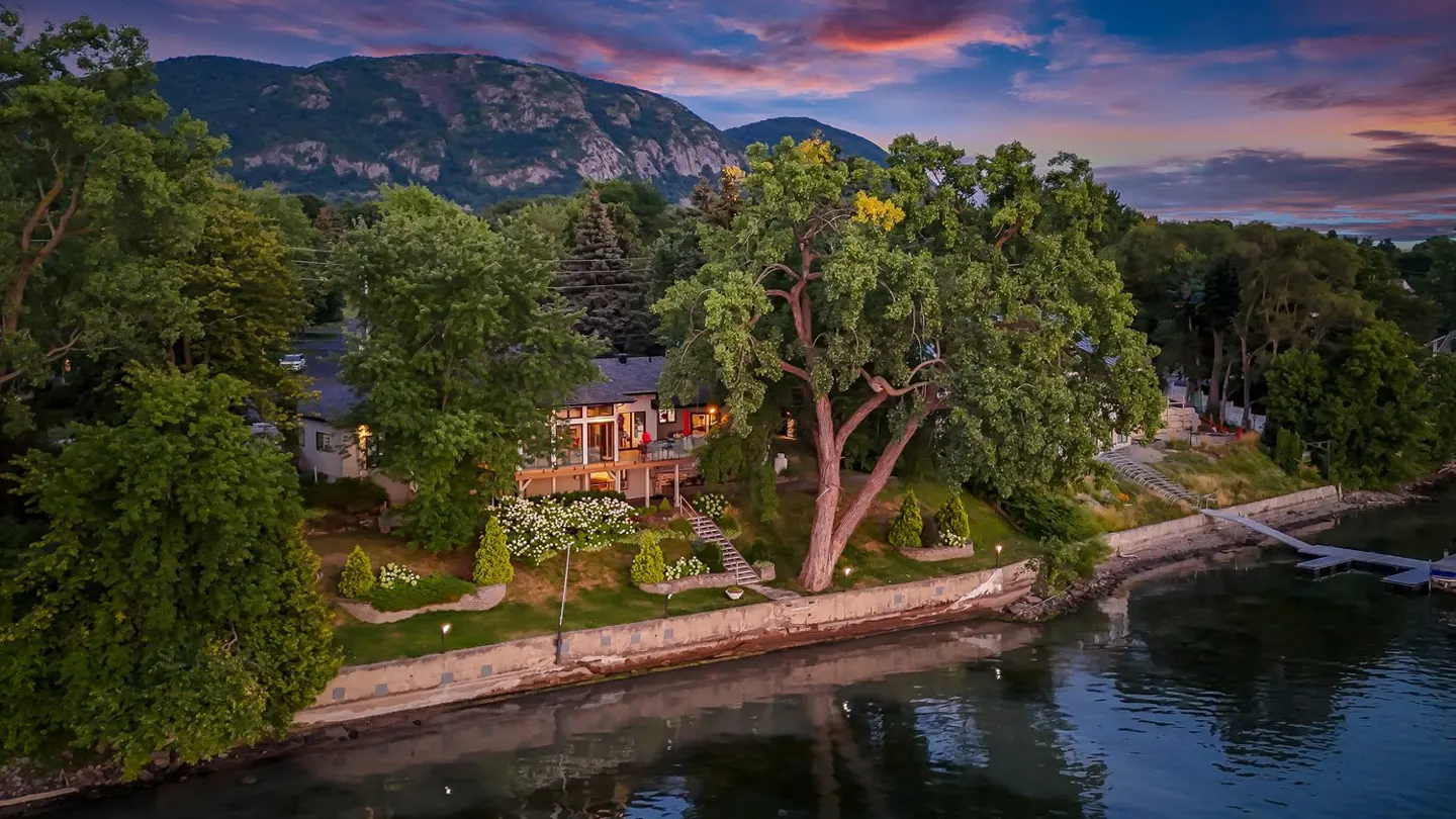 Waterfront home with a deck, surrounded by trees, with a mountain backdrop under a colorful sunset sky.