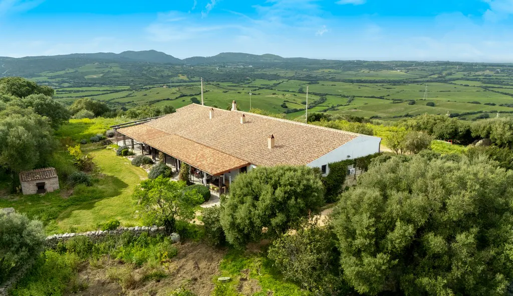 Aerial view of a large, single-story house with a brown tile roof, surrounded by green trees and a rolling green landscape under a blue sky.