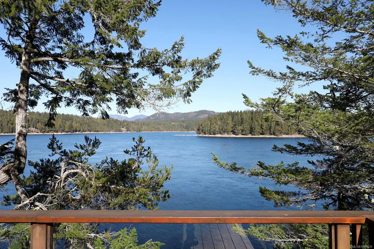 A scenic view of a blue lake framed by green trees and a wooden railing under a clear blue sky.