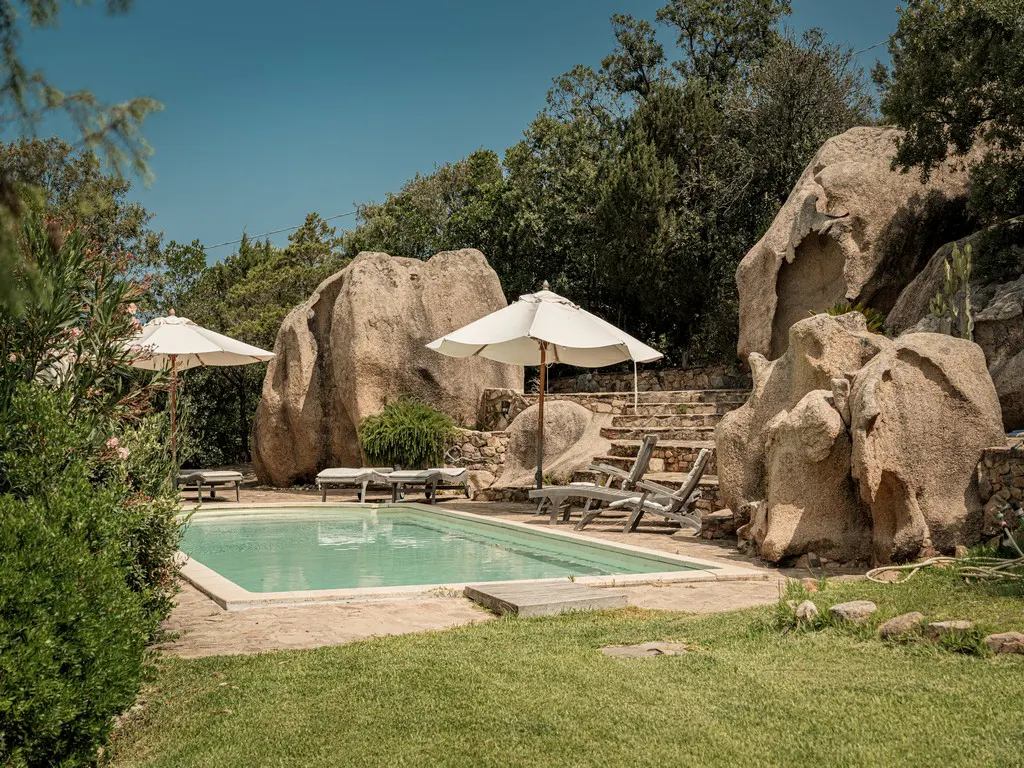 Outdoor pool area with lounge chairs, white umbrellas, and large rock formations. Green grass and trees surround the pool.