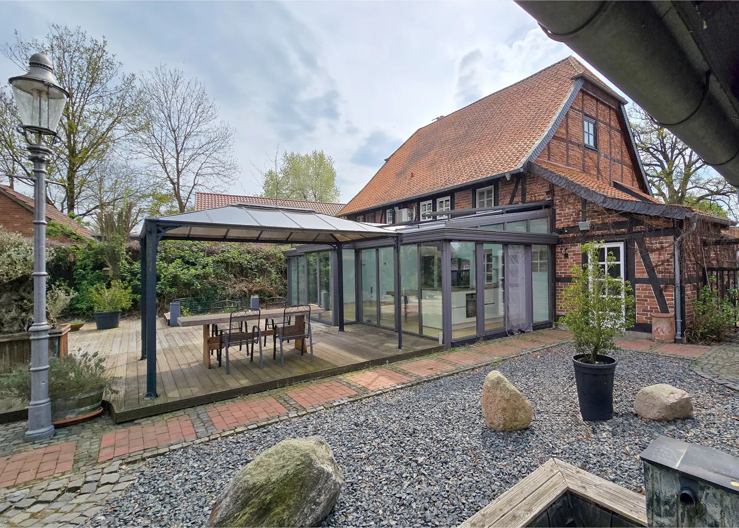Outdoor patio with a wooden deck, table, chairs, and a glass-enclosed sunroom attached to a brick house with a red tile roof.