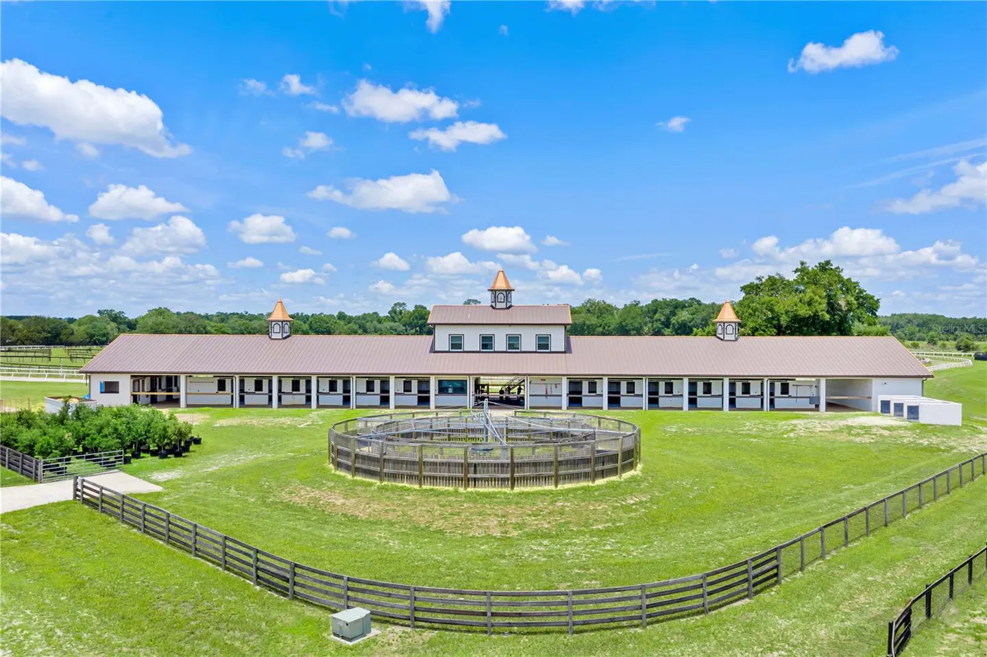 Aerial view of a horse barn with a brown roof, white walls, and a round pen in a green field under a blue sky.