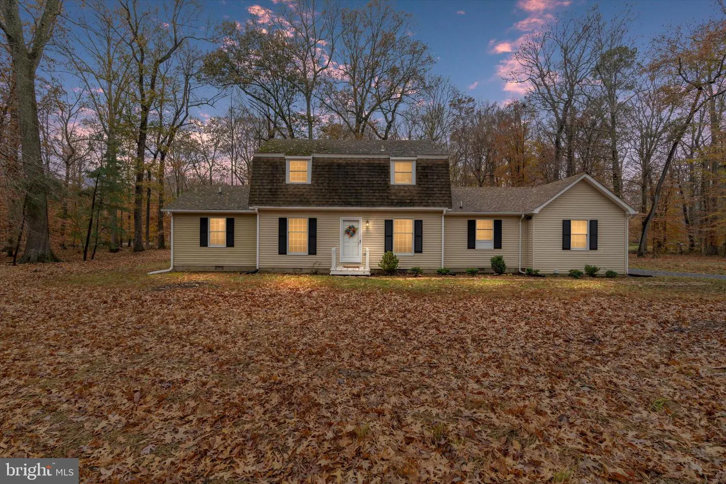 Beige house with black shutters and a white door with a wreath, surrounded by trees and a yard covered in fallen leaves.
