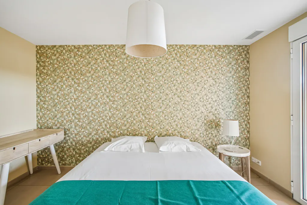 Bedroom with geometric wallpaper, white bed with teal blanket, and light wood desk.