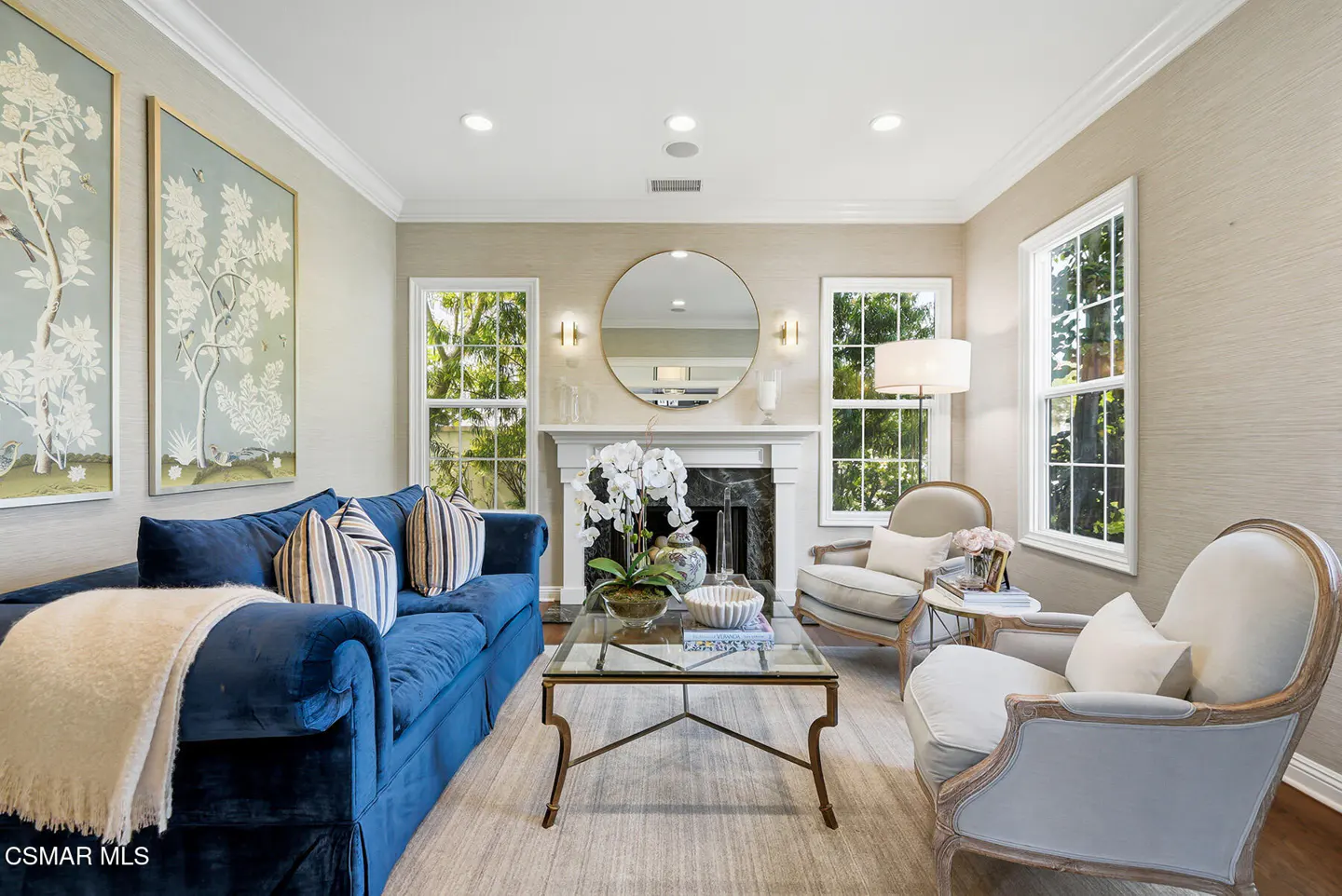 Living room with blue sofa, two armchairs, glass coffee table, fireplace, and round mirror. Two large paintings on the wall.