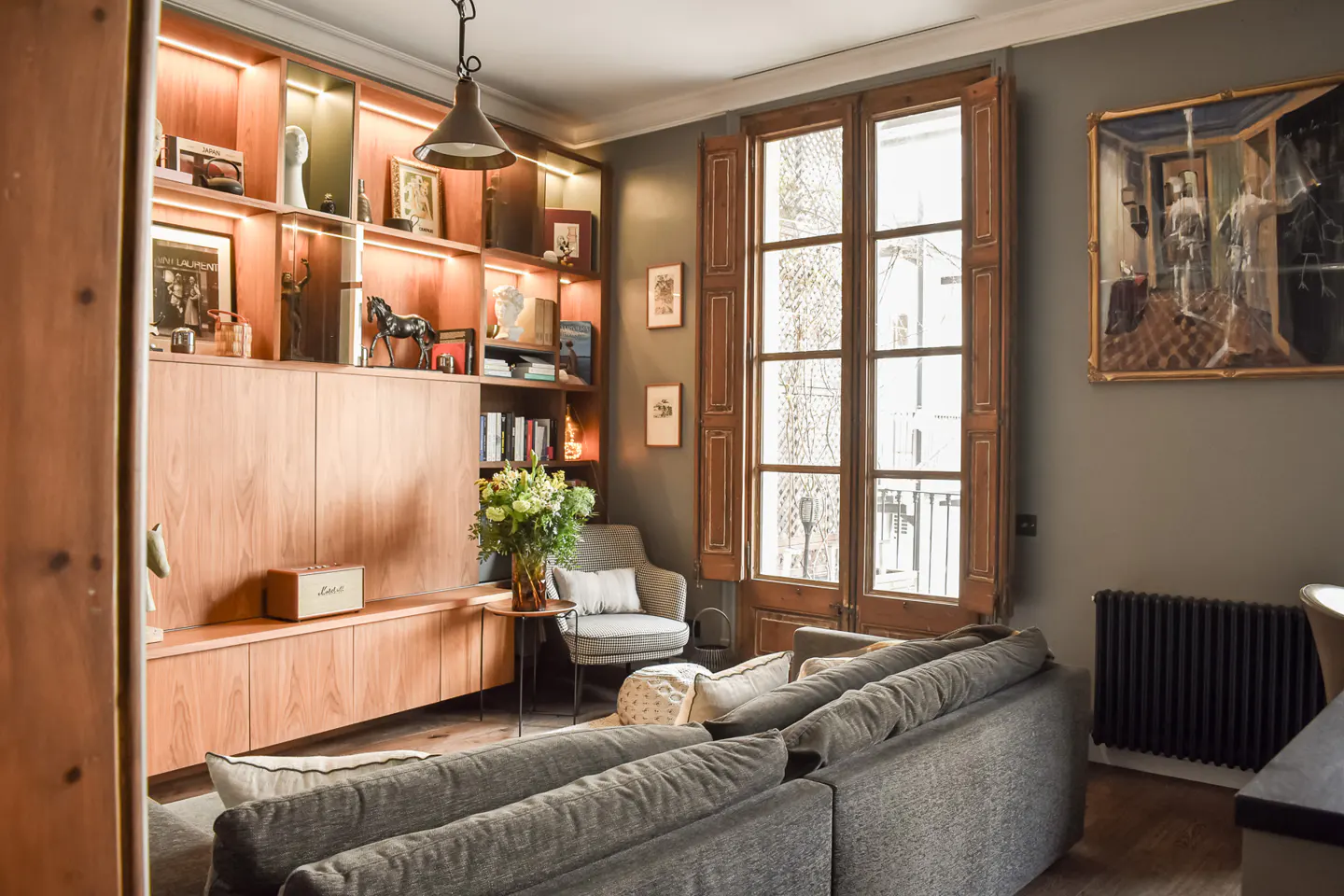 Living room with gray sofa, wooden built-in shelves with books and decor, and a window with wooden shutters.