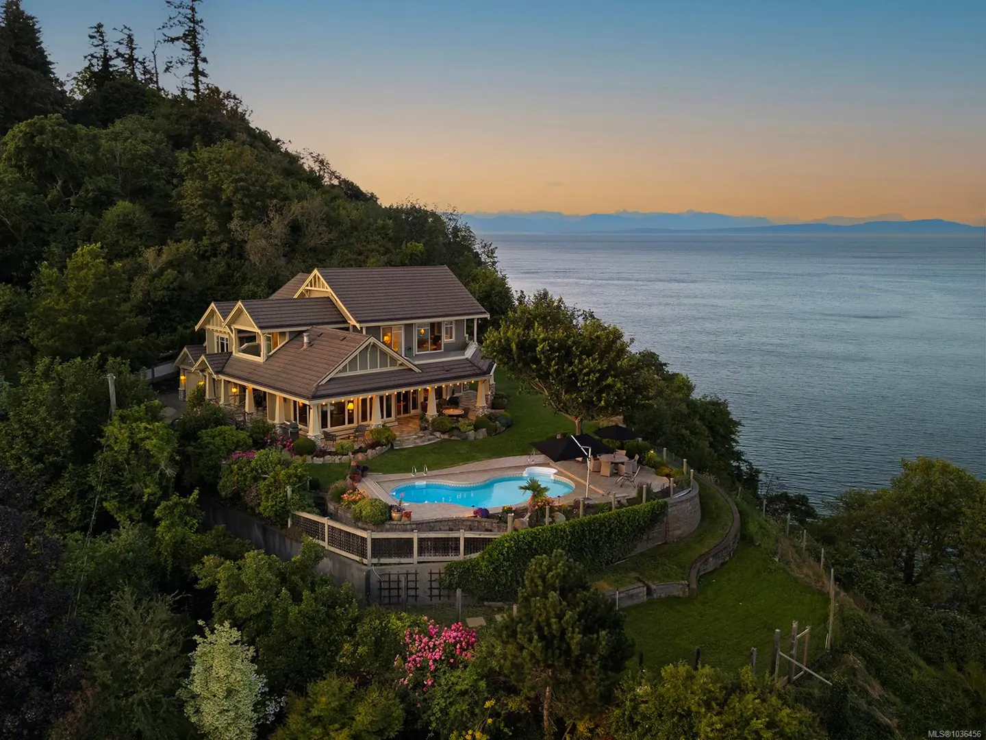 Aerial view of a large, multi-story house with a pool overlooking the ocean at sunset. Lush greenery surrounds the property.