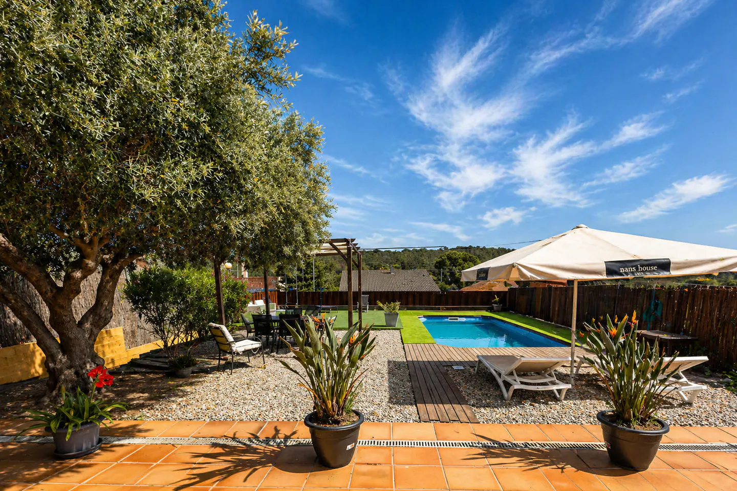 Outdoor pool area with lounge chairs, umbrella, and dining set. A large tree provides shade. Blue sky with clouds above.