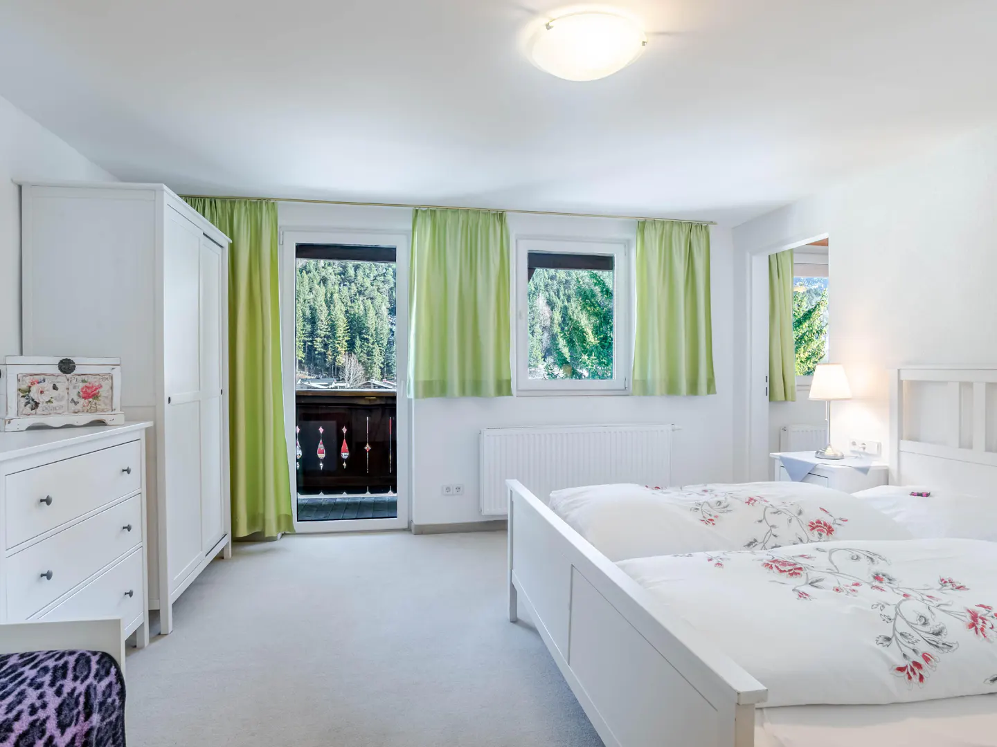 Bright bedroom with two beds, white furniture, and green curtains. A balcony overlooks a forest.