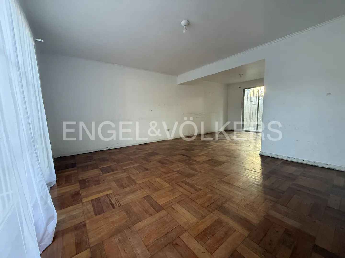 Empty room with parquet floor, white walls, and Engel & Völkers logo. Natural light streams in from a window.
