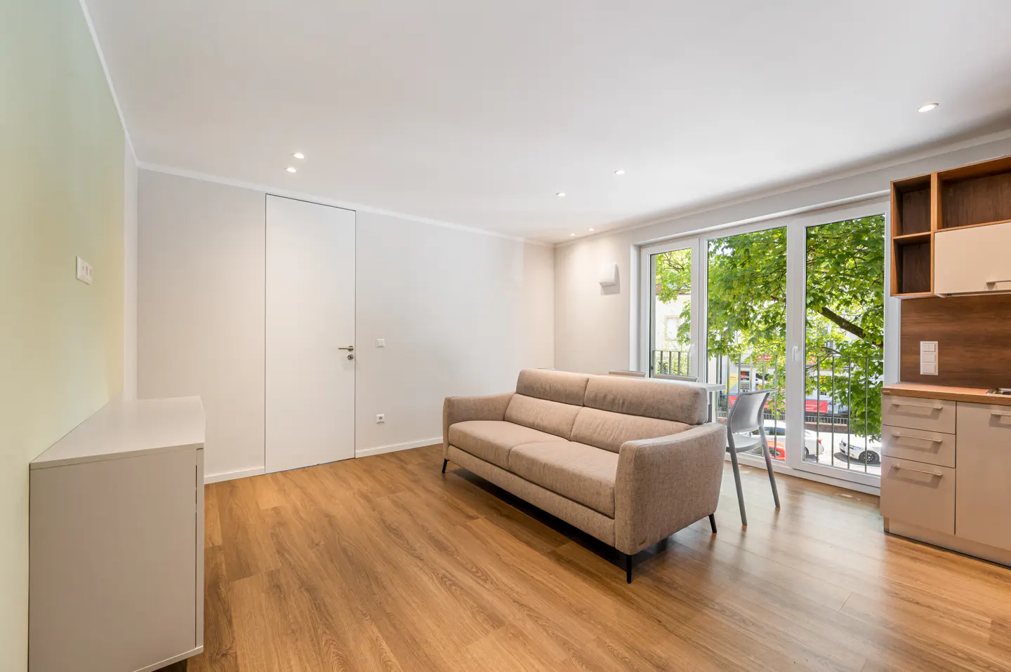 Bright apartment interior with wood floors, beige sofa, white walls, and a sliding glass door to a balcony with green trees.