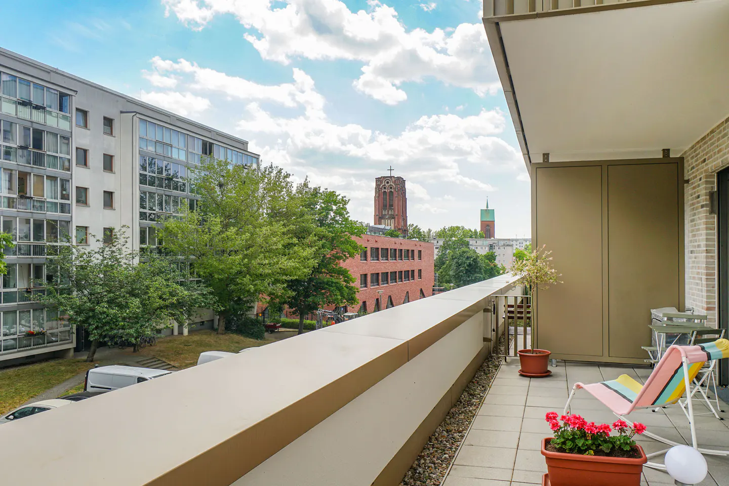 Balcony view with a colorful striped lounge chair, potted plants, and a cityscape featuring a church tower in the background.