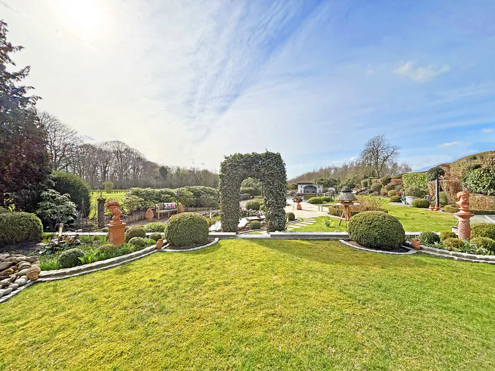 A wide shot of a green lawn with an ivy archway, bushes, and trees under a blue sky.