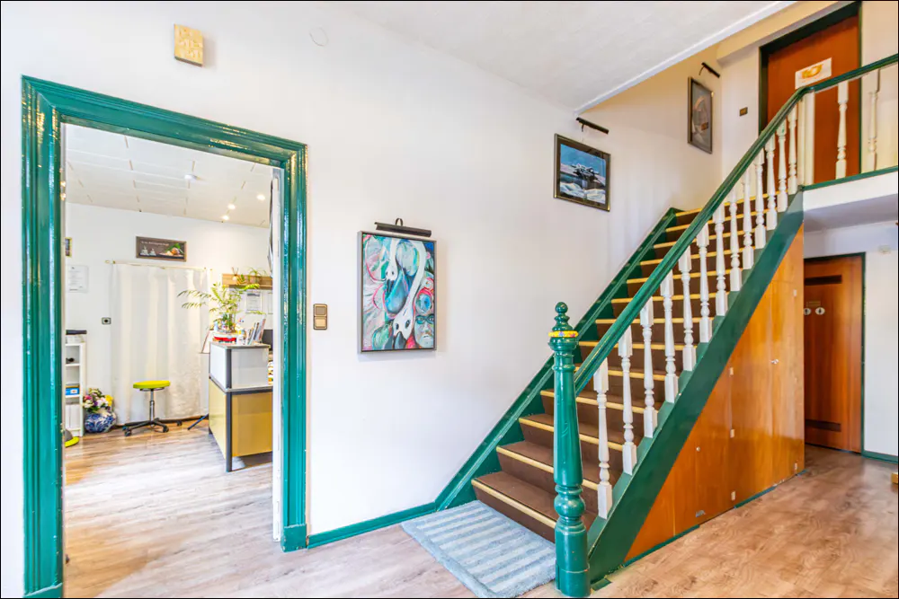Interior view of a building with a green-framed doorway, stairs with green railings, and artwork on white walls.
