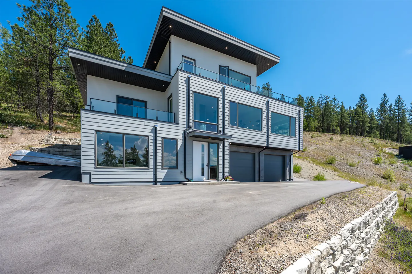 Modern three-story home with gray siding, large windows, balconies, and a two-car garage on a hillside with pine trees.