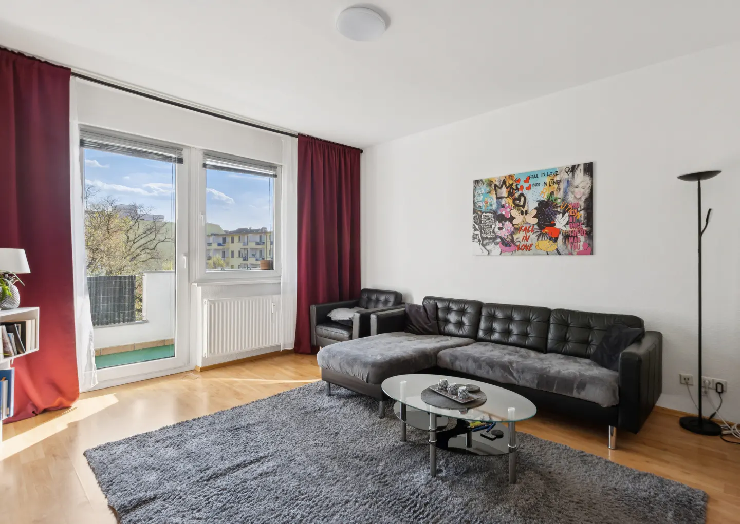 Living room with a black sectional sofa, gray rug, glass table, and red curtains. Balcony view through the window.