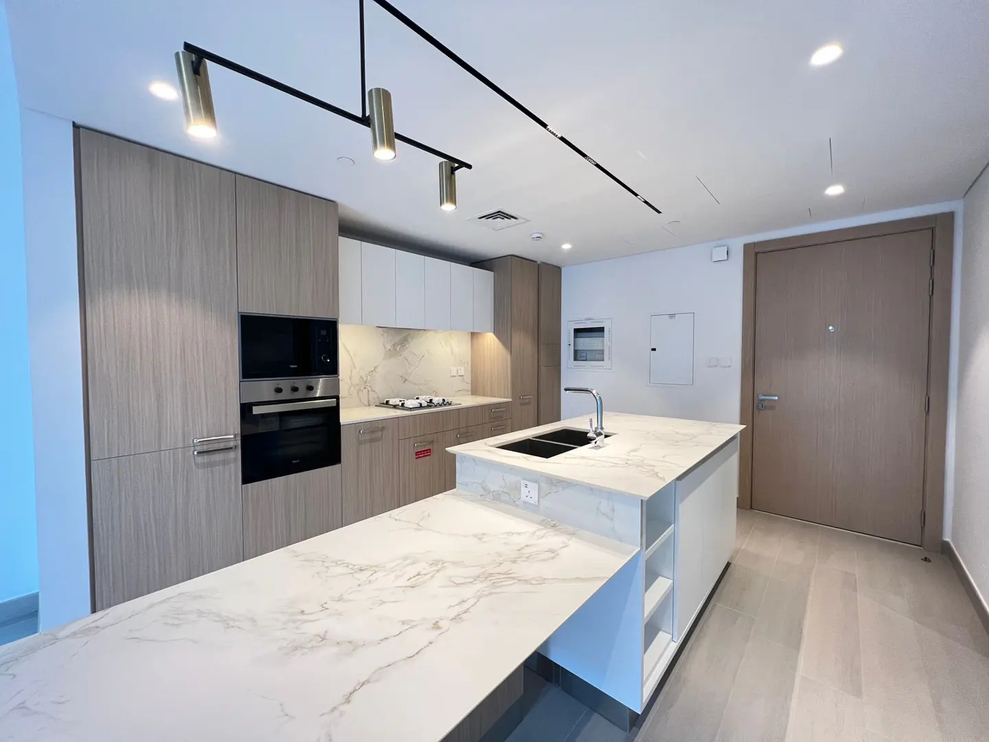 A modern kitchen with light wood cabinets, marble countertops, and stainless steel appliances. A large island with a sink is in the center.