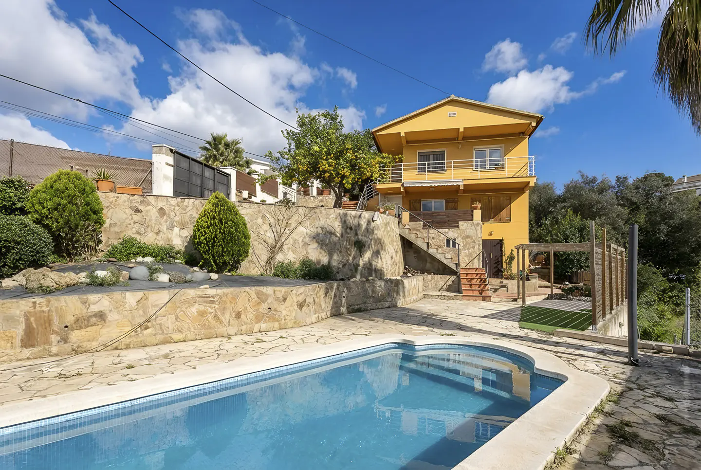 Two-story yellow house with a blue swimming pool in the foreground on a sunny day.