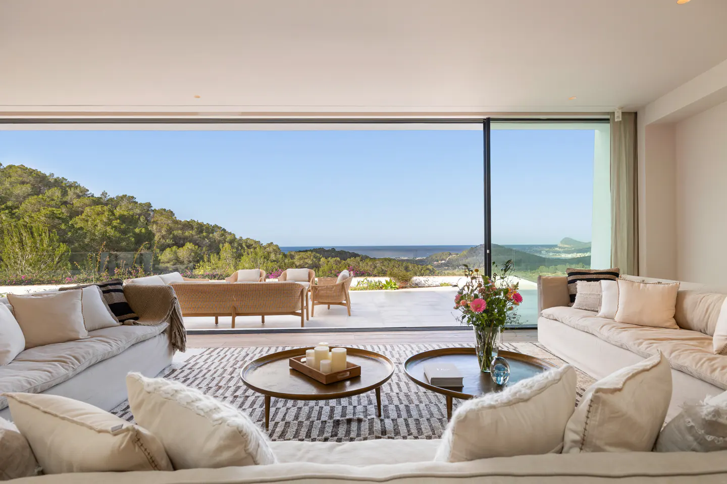 Bright living room with white sofas, round tables, and a view of the ocean and green hills through a large sliding glass door.
