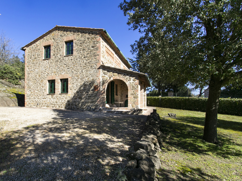 A two-story stone house with green-trimmed windows and an arched entryway, set on a gravel lot beside a large tree.