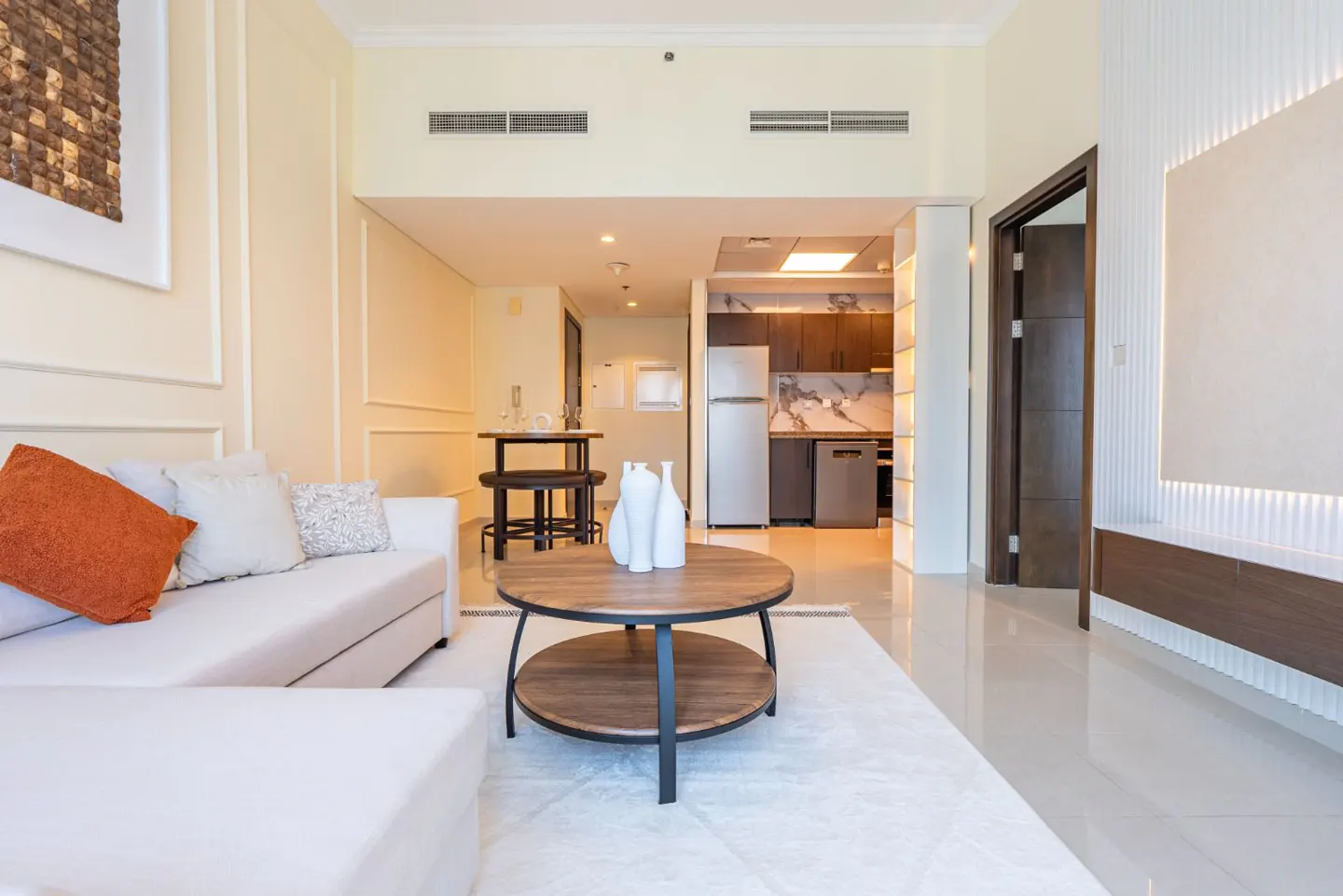 Bright, modern apartment interior with a white sofa, round wood coffee table, and open kitchen with stainless steel appliances.