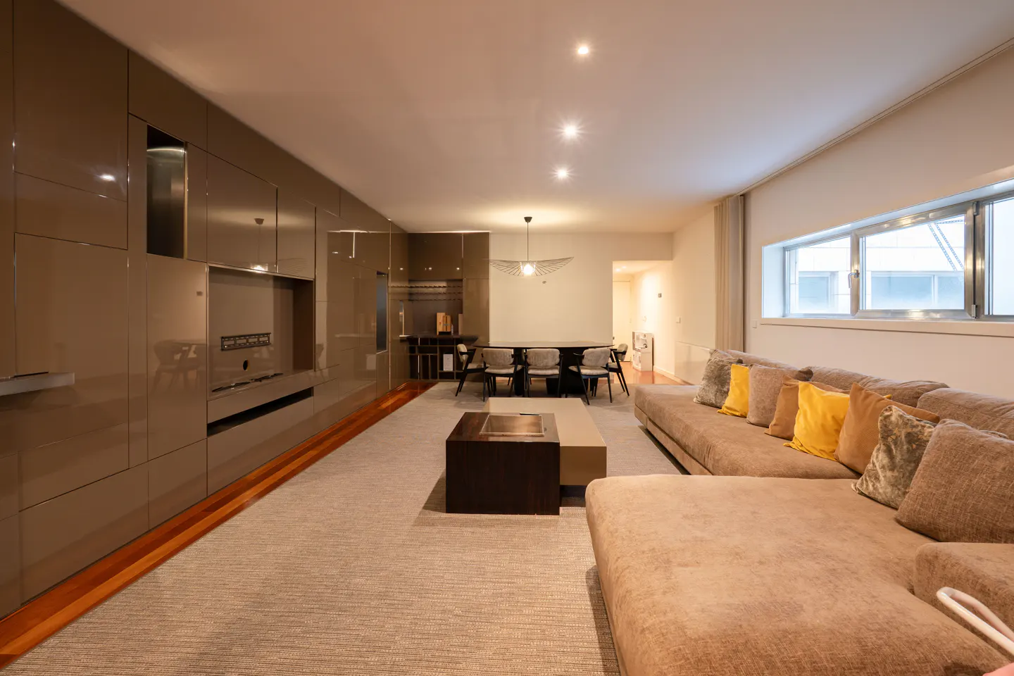 A modern living room with a brown sectional sofa, a dark wood coffee table, and a dining area in the background.