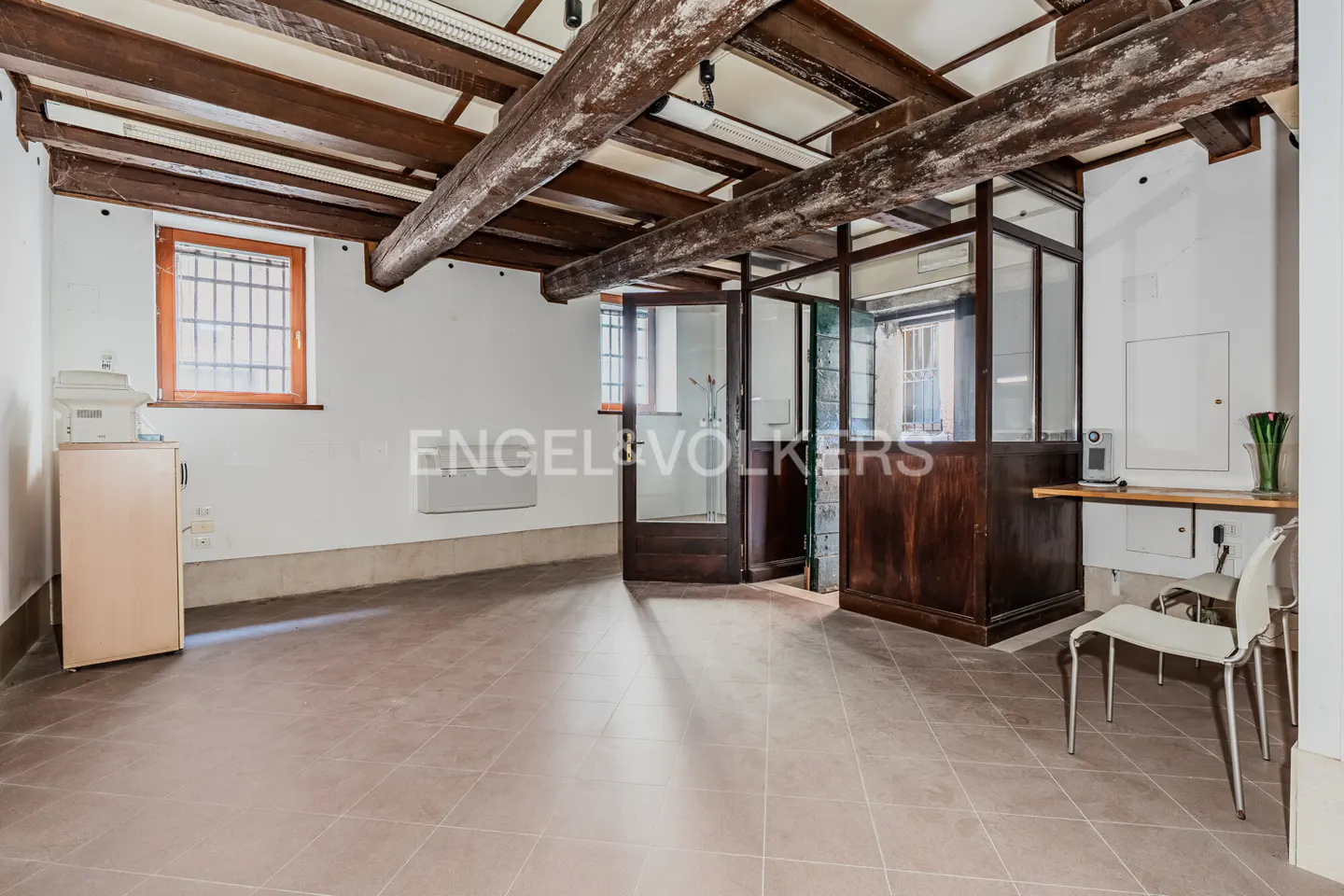 Interior view of a room with exposed wooden beams, tile flooring, and a glass-enclosed area. Two white chairs are near a small counter.
