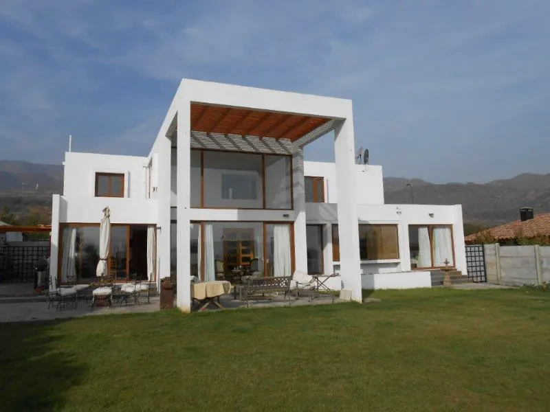 Modern white house with large windows and a pergola, set on a green lawn with mountains in the background. Outdoor furniture is visible.