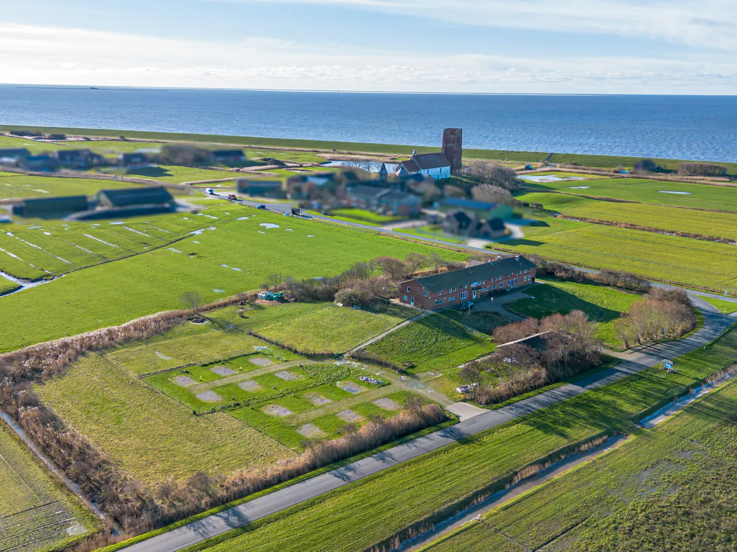 Aerial view of a long brick building in a rural green landscape near the coast, with a distant church tower by the sea.
