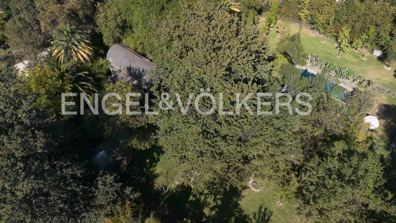 Aerial view of a house with a gray roof surrounded by green trees and a pool. Engel & Völkers logo is superimposed.
