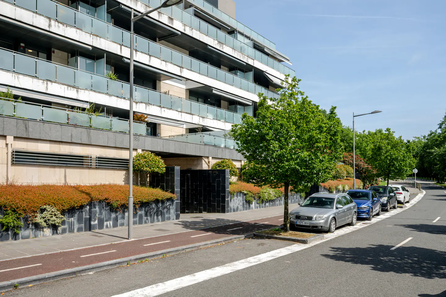 Modern apartment building with glass balconies, street with parked cars, and green trees under a blue sky.