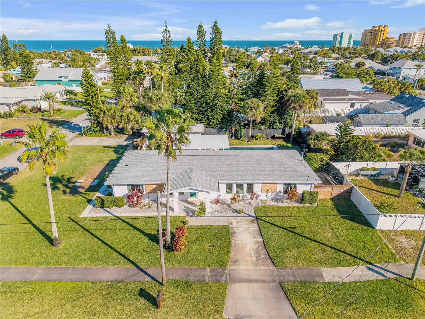 Aerial view of a white ranch-style house with a gray roof, surrounded by green lawn, palm trees, and other houses near the ocean.