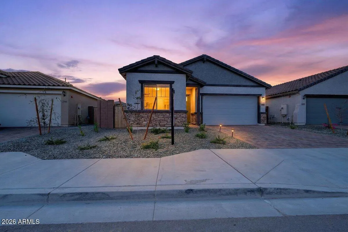 A single-story home with a gray exterior and a two-car garage at dusk. The sky is pink and purple. The yard is landscaped with rocks and small plants.