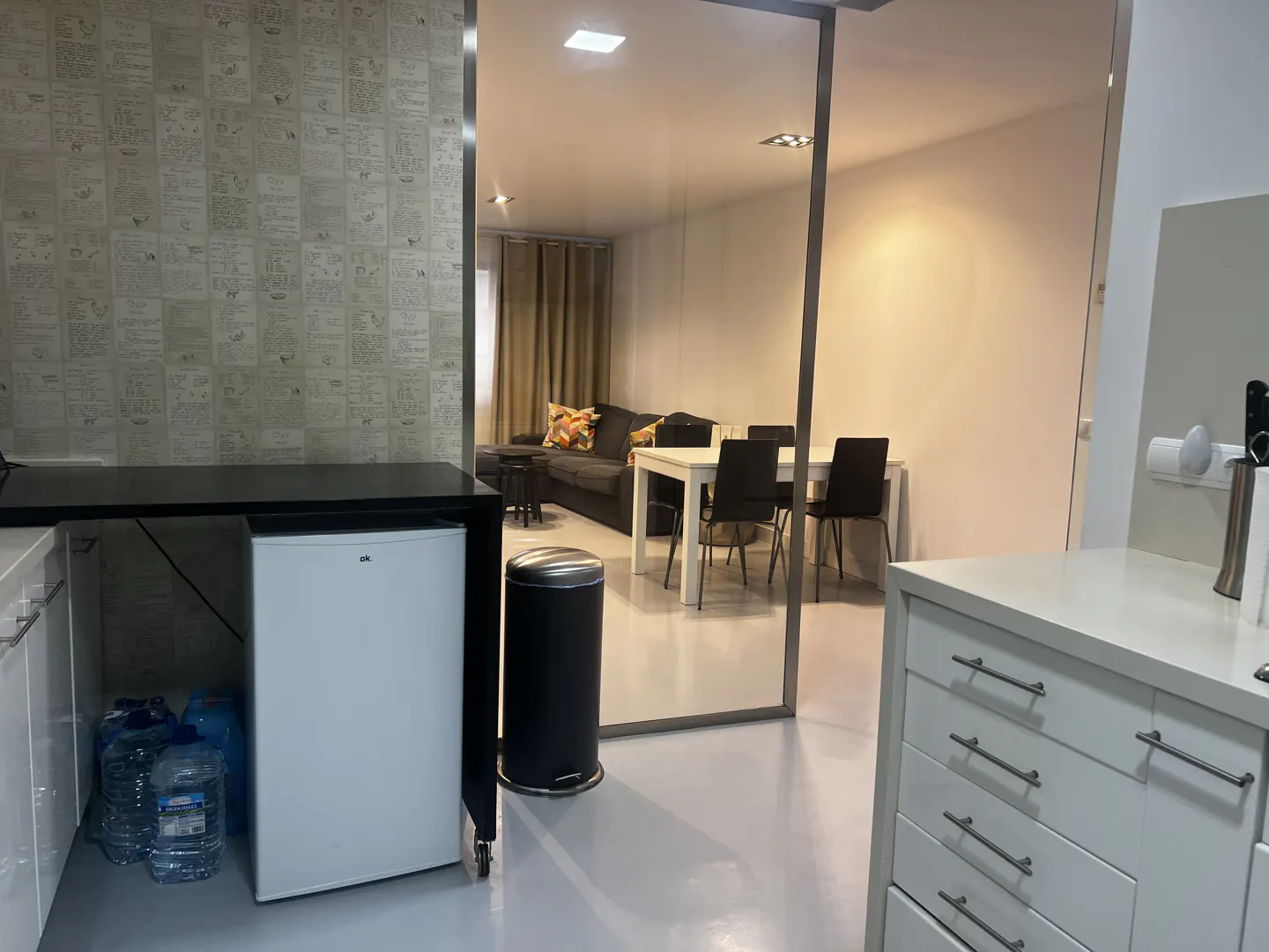 A modern apartment interior with a white kitchen, a mini-fridge, and a view of the living room with a gray sofa and dining table.