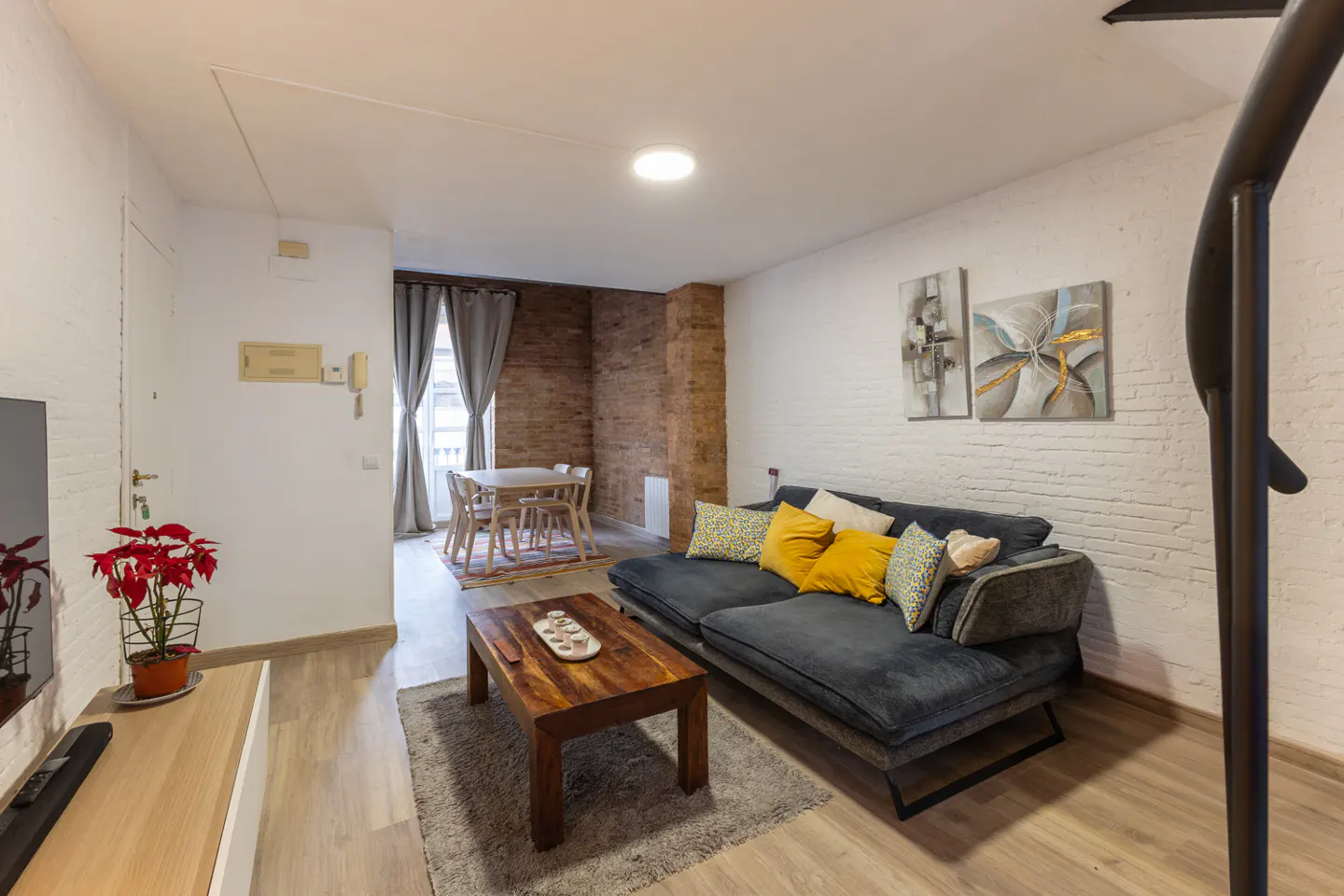 Living room with gray sofa, yellow pillows, wood coffee table, and dining area. Brick and white walls, art, and spiral staircase.
