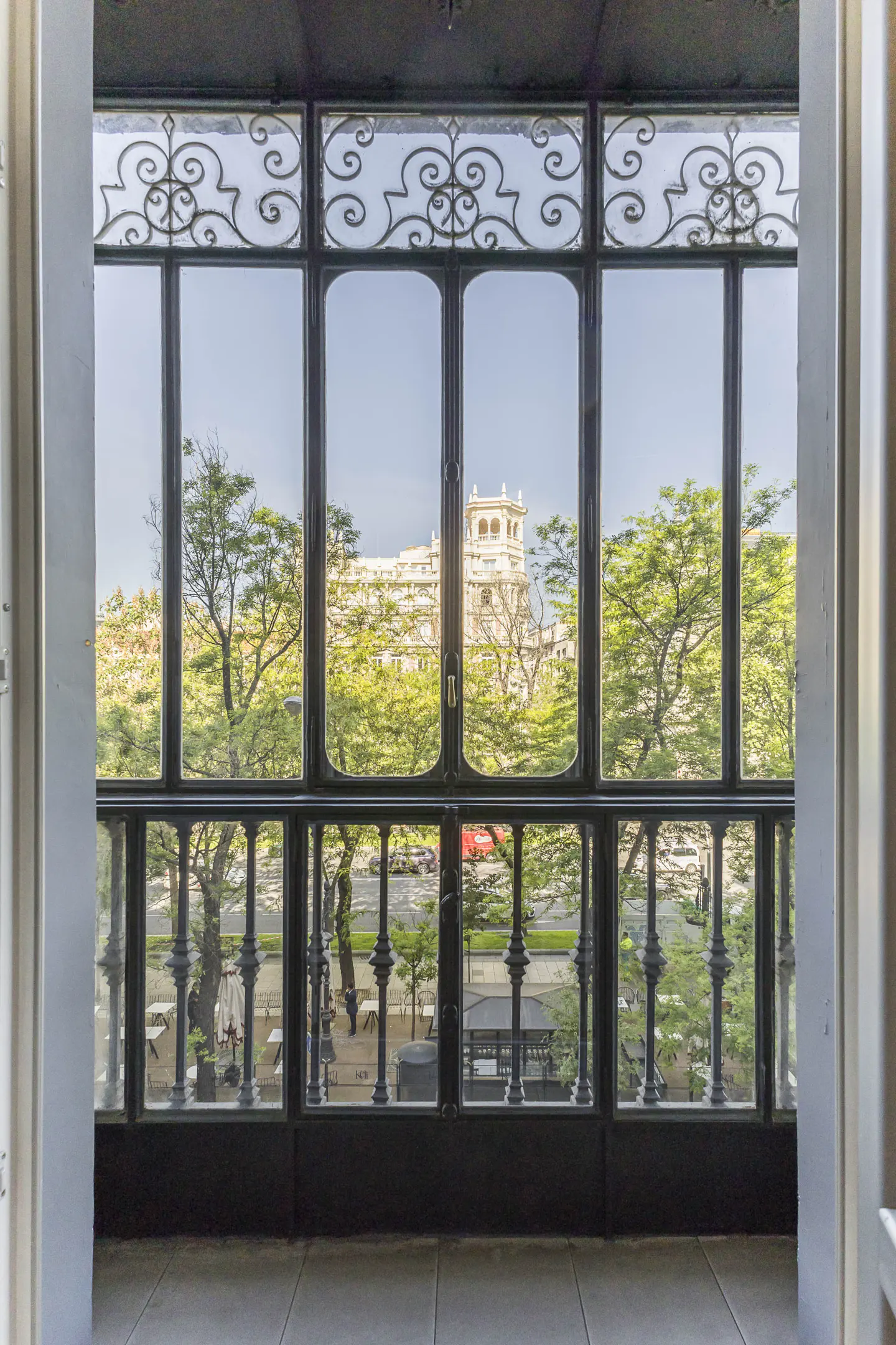 View from a balcony with black ironwork, showing trees, a street, and a building in the distance.