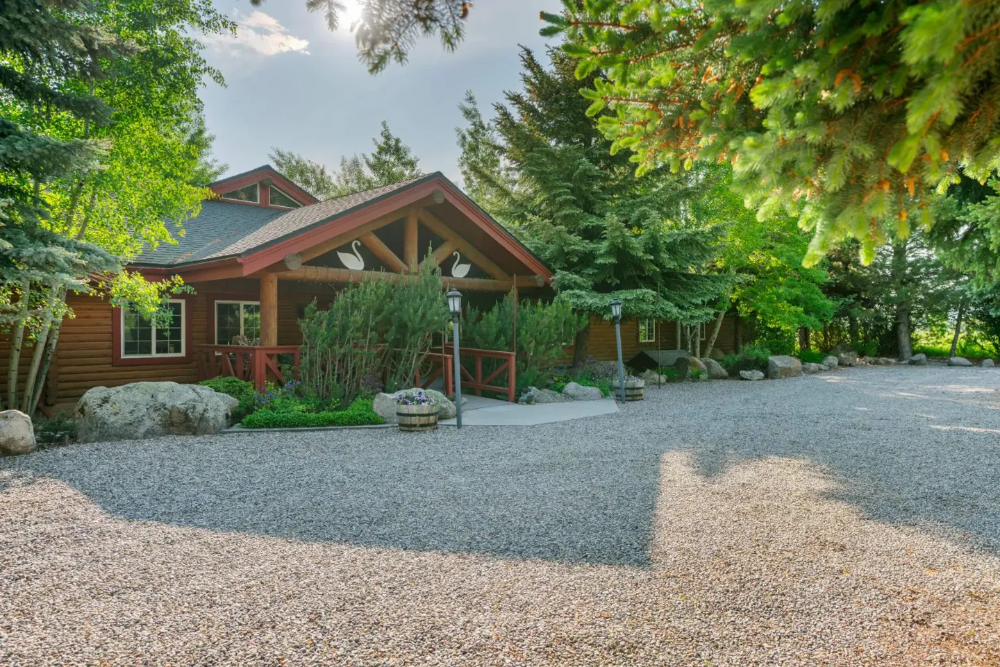 A rustic log cabin with a gravel driveway, surrounded by lush green trees and decorated with swan motifs on the gable.