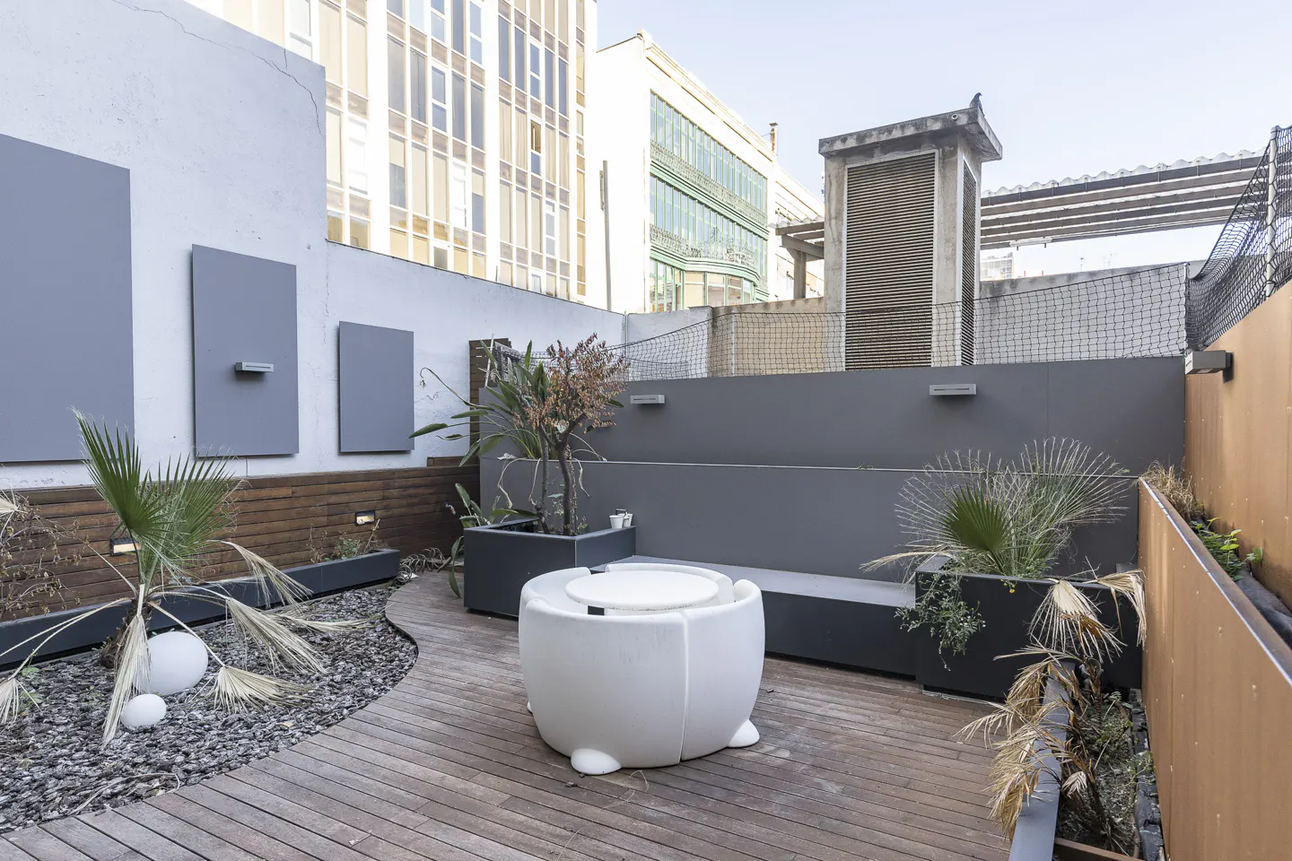 Modern rooftop terrace with wooden decking, a white circular table set, potted plants, and city buildings in the background.
