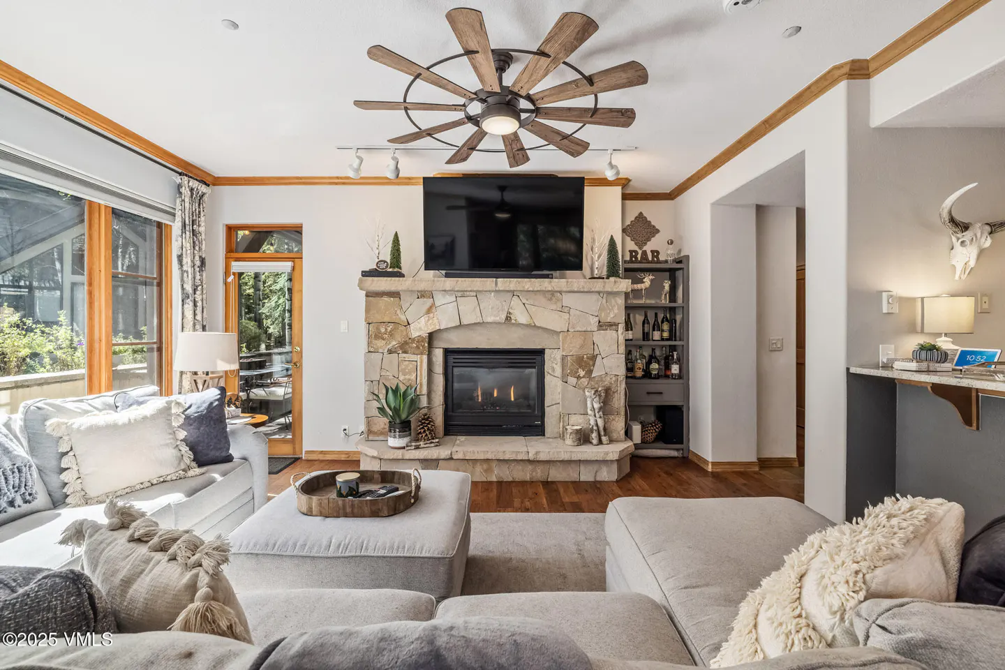 Living room with stone fireplace, TV, and large ceiling fan. Gray sectional sofa with decorative pillows. Bar area with wine rack.