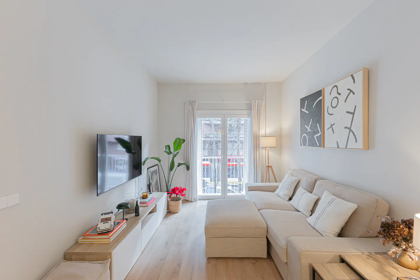 Bright living room with a beige sectional sofa, TV, and balcony. Abstract art hangs on the white wall. A plant adds a touch of green.