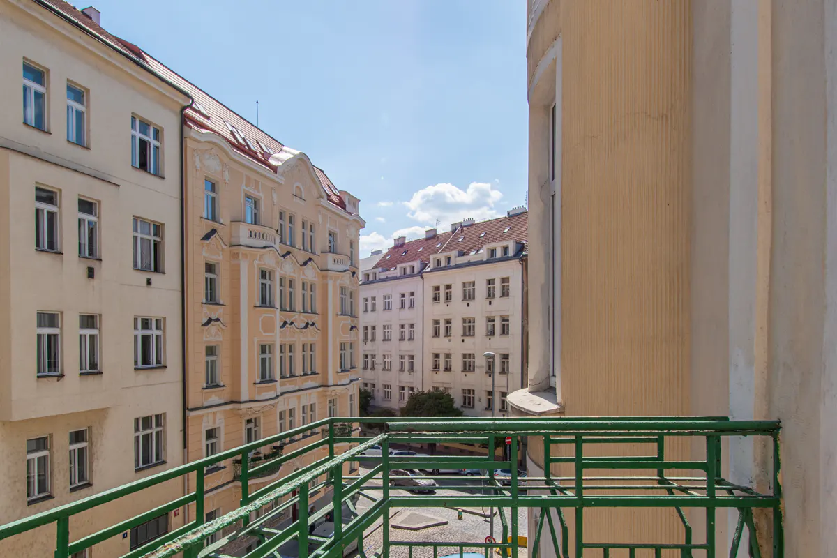 View from a green balcony overlooking a European city street with beige buildings and red tile roofs under a blue sky.