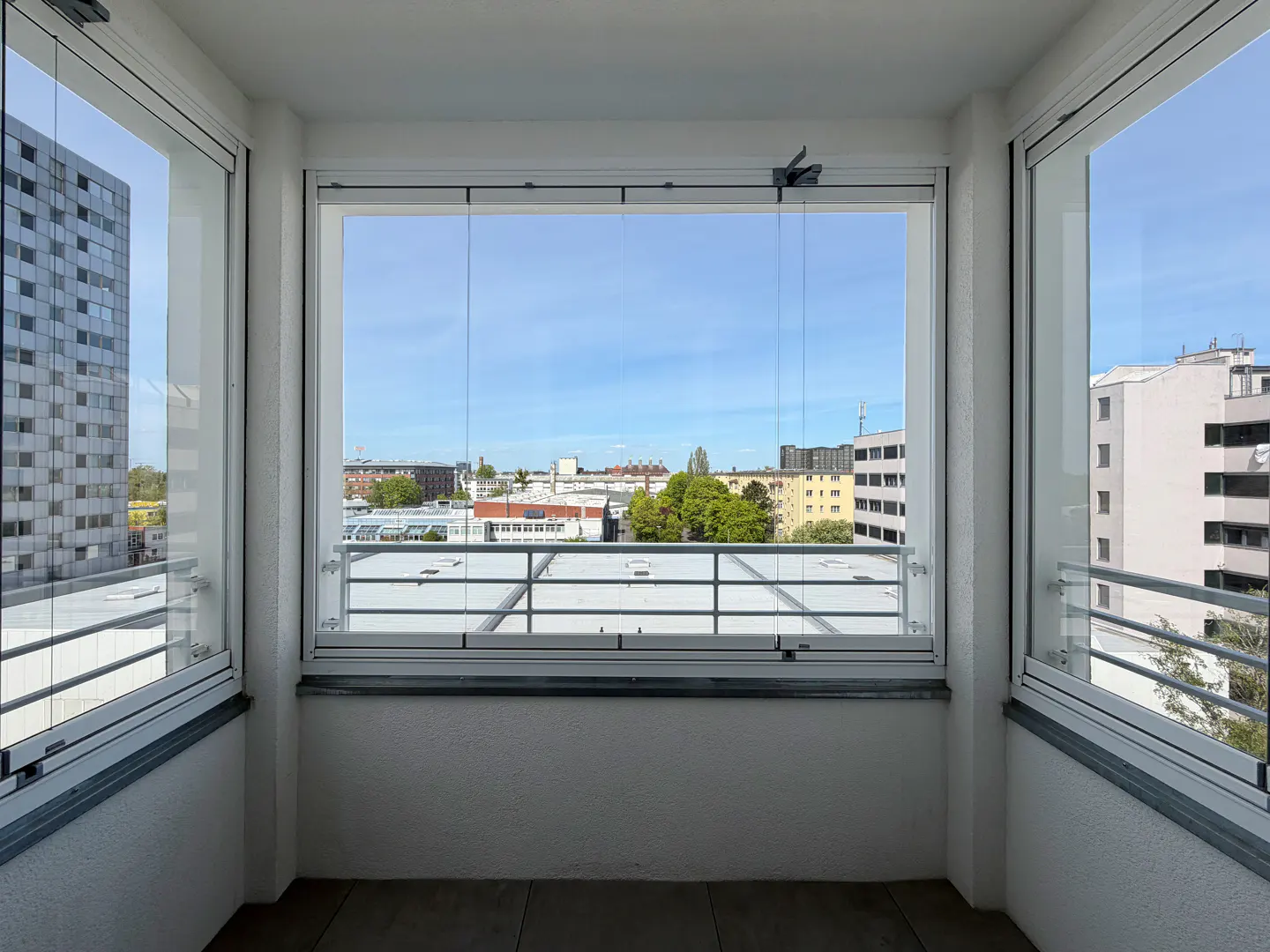 Balcony view with clear glass panels framing a cityscape under a blue sky. White walls and gray trim add a modern touch.