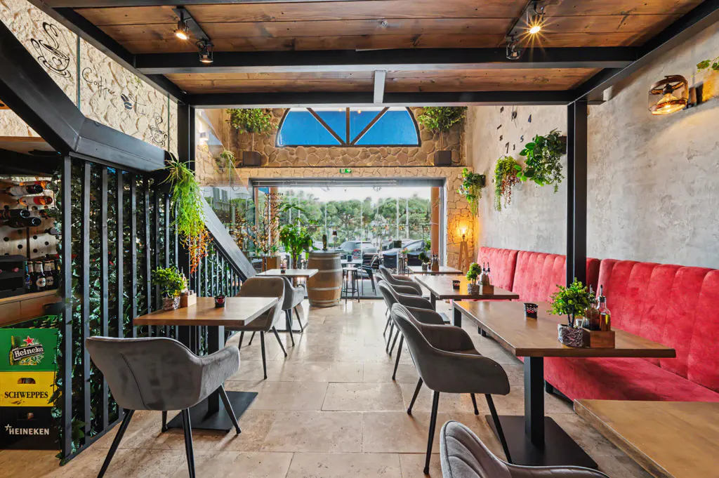 Restaurant interior with wooden tables, gray chairs, and a red cushioned bench. Stone walls, plants, and a barrel add to the ambiance.