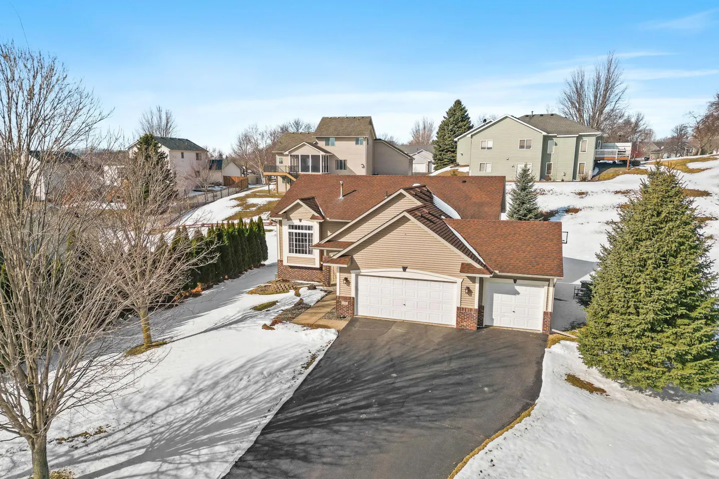 Aerial view of a tan two-story house with a brown roof, a two-car garage, and a black driveway in a snowy neighborhood.