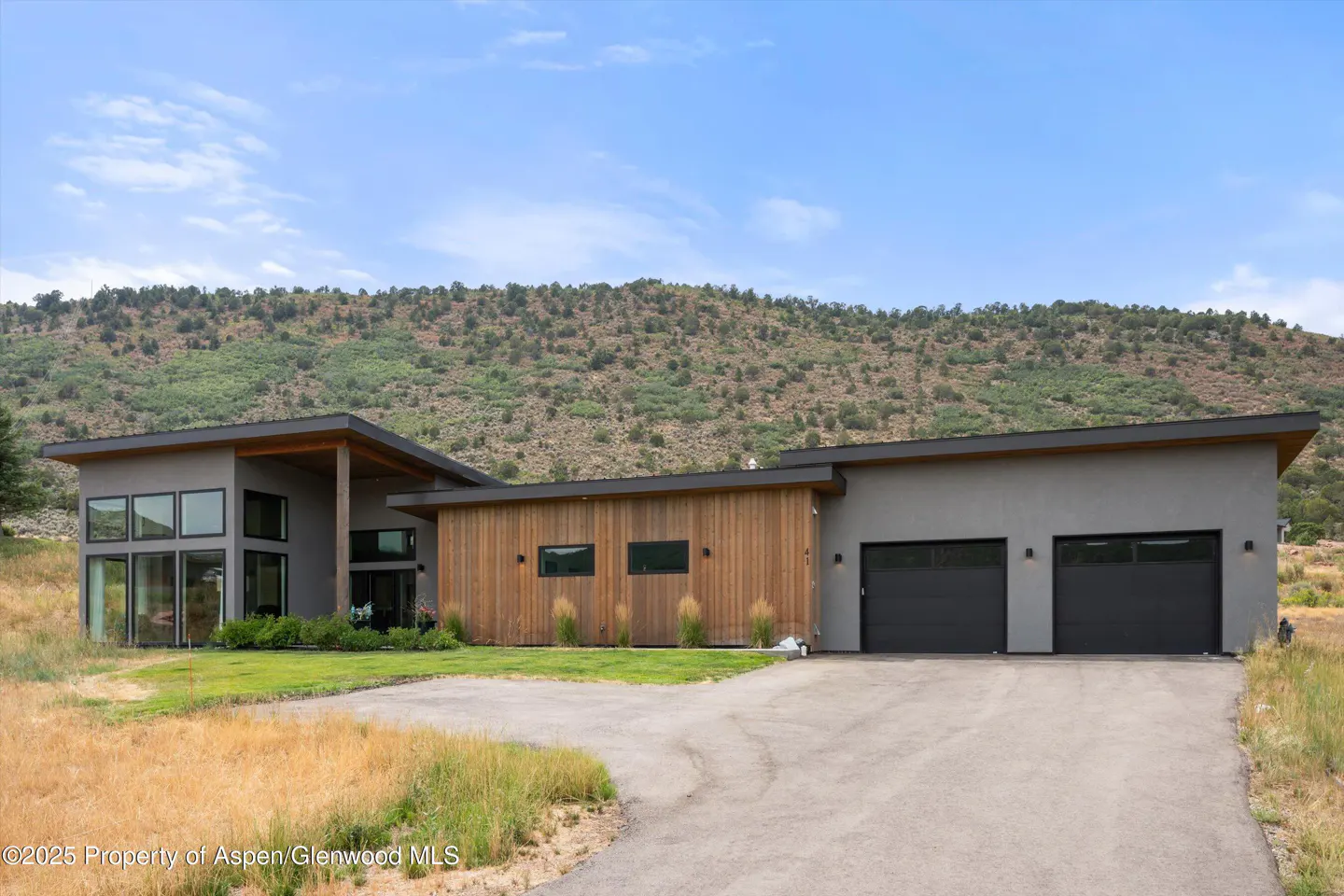 Modern home with gray stucco and wood siding, black garage doors, and a mountain backdrop.