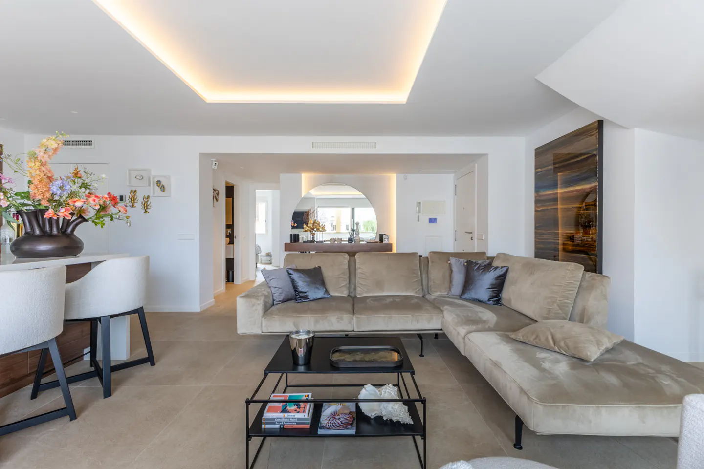 Bright living room with beige sectional sofa, black coffee table, and bar with flowers. White walls and recessed lighting.