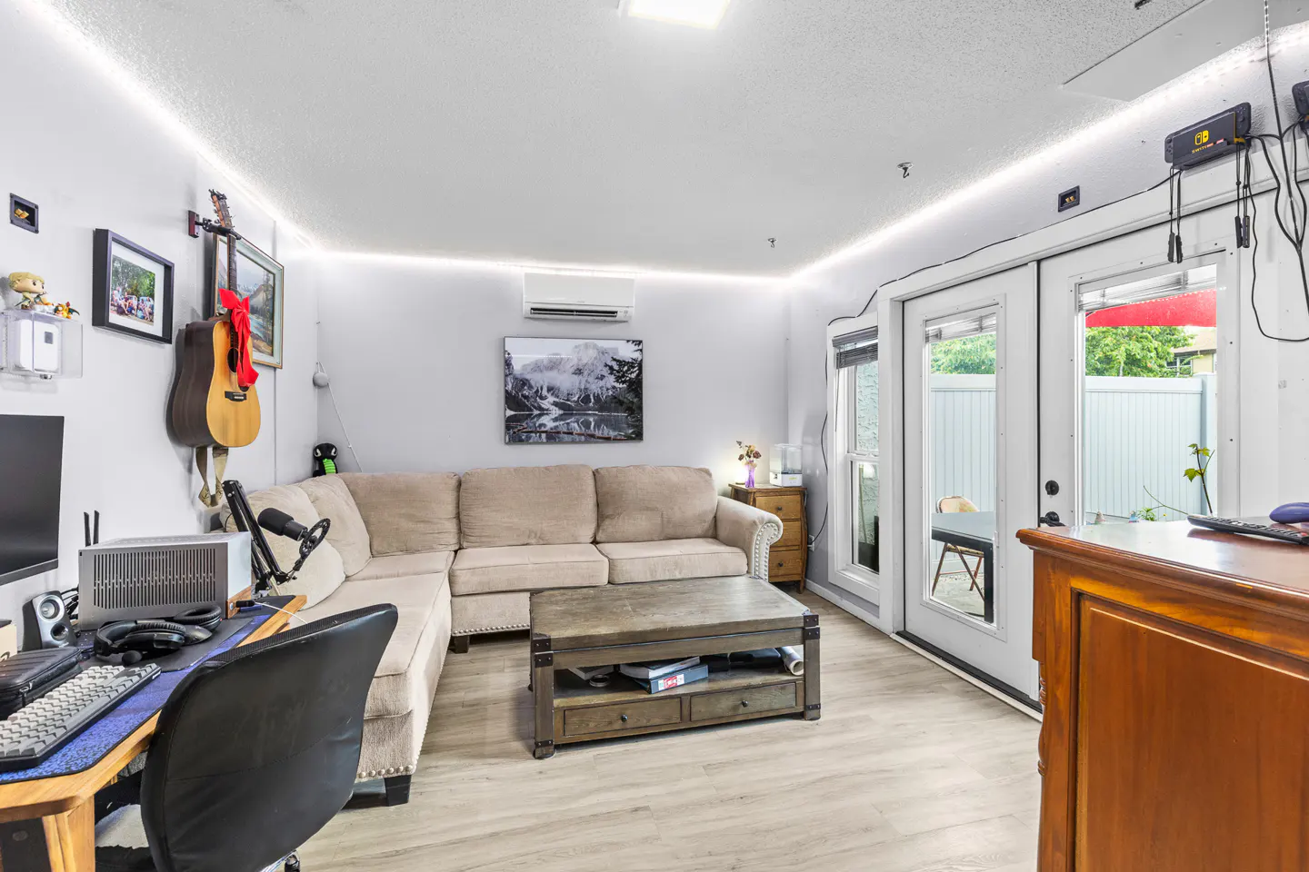A cozy living room with a beige sectional sofa, wooden coffee table, desk setup, and glass doors leading to a patio.